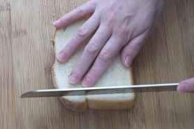 One hand on top of a slice of white bread with the other hand using a knife to cut the crust from the side of the bread.