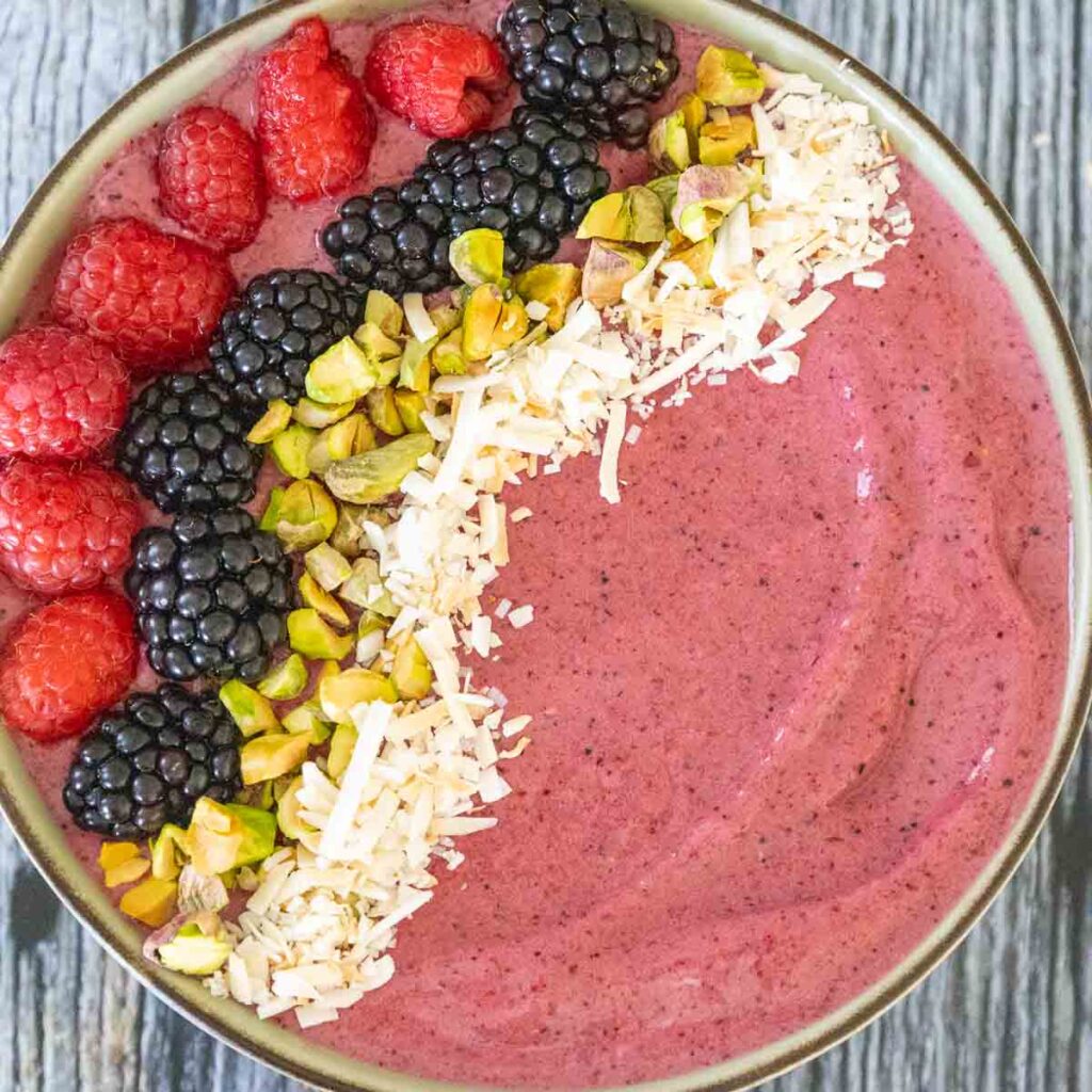 A green bowl with a brown rim filled with a mixed berry pinkish red smoothie and topped from left to right with a line of red raspberries, blackberries, chopped green pistachios, and toasted coconut.