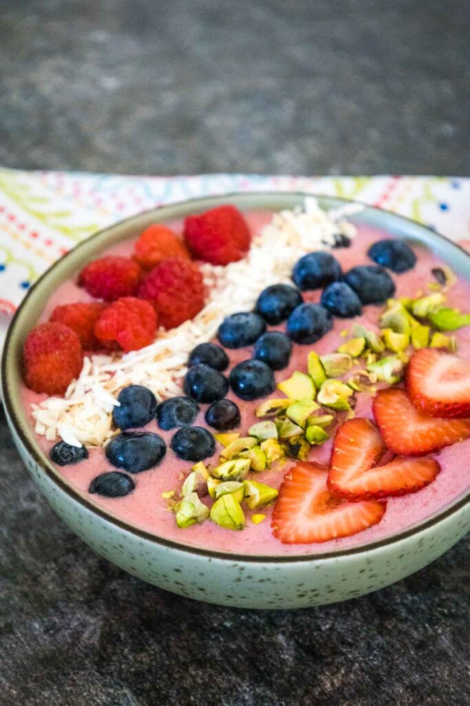 light pink smoothie bowl topped with raspberries, coconut, blueberries, pistachios, and strawberry slices.