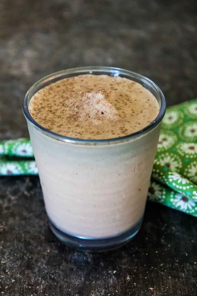 A glass filled with chocolate tofu smoothie sprinkled on the top with grated chocolate with a green and white flowered napkin blurred in the background.