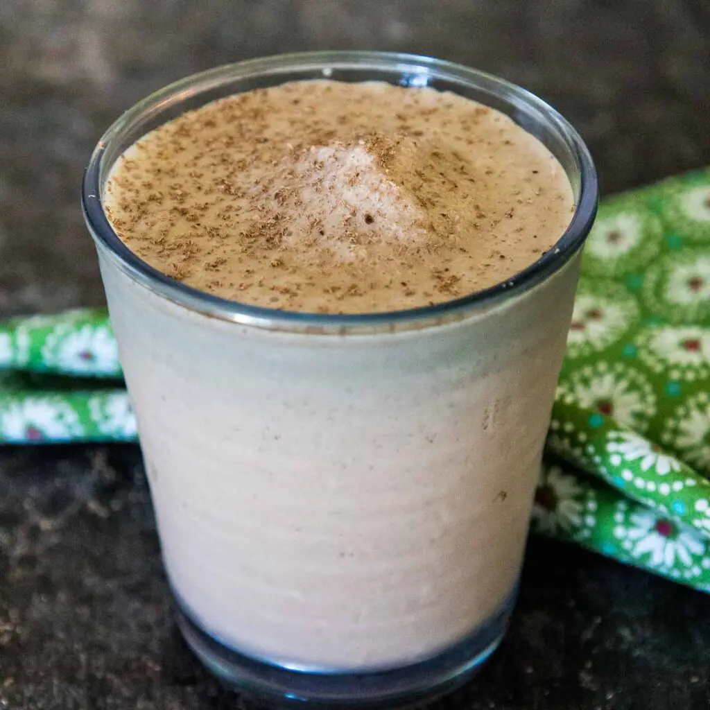 A glass filled with chocolate tofu smoothie sprinkled on the top with grated chocolate with a green and white flowered napkin blurred in the background.