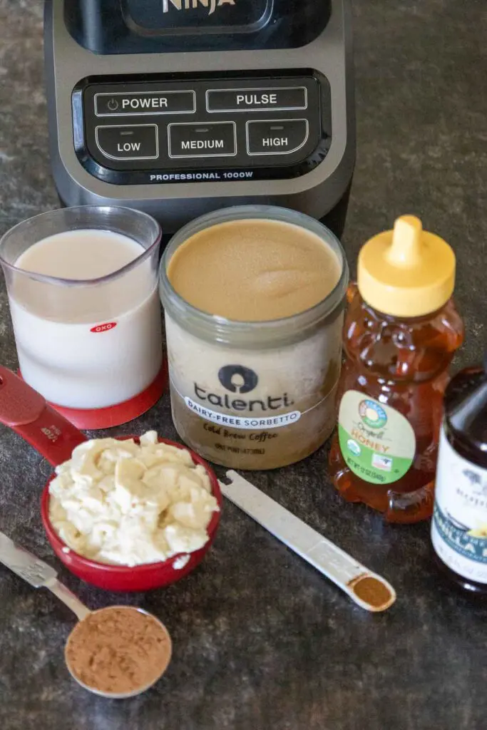 From back to front, bottom of blender with almond milk, dairy free sorbet, honey, vanilla extract in front, with tofu in a red measuring cup, cinnamon in a white measuring spoon and cocoa powder in a silver measuring spoon in the front.