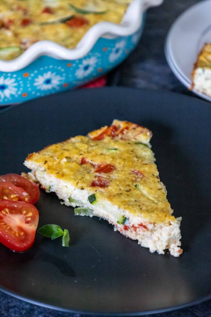 One slice of impossible zucchini pie on a black plate with a cherry tomato cut into 3 slices to the left as garnish with the remaining pie blurred in the background.