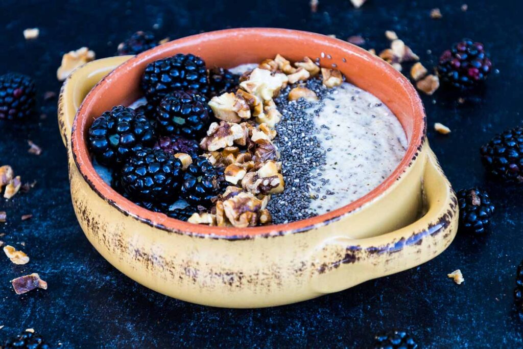 Overhead and up close shot of Maple Walnut Noatmeal in a tan and orange bowl and a black backdrop with blackberries arranged on the top edge of the bowl, followed with toasted walnuts and chia seeds with blackberries and toasted walnuts scattered around the outside of the bowl.