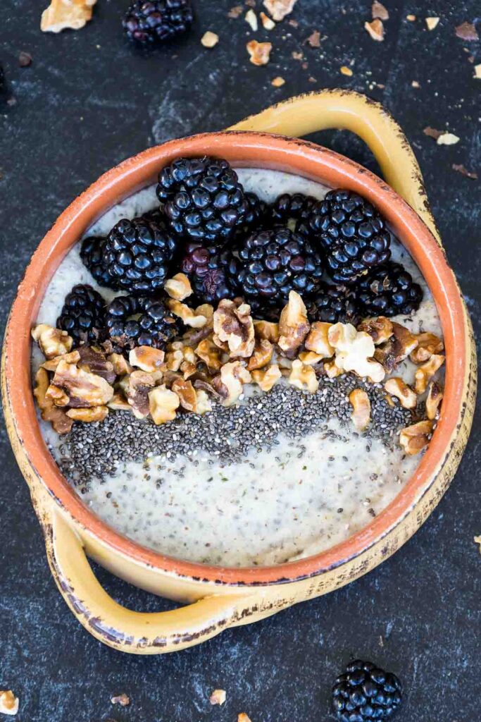 Overhead and up close shot of Maple Walnut Noatmeal in a tan and orange bowl and a black backdrop with blackberries arranged on the top edge of the bowl, followed with toasted walnuts and chia seeds.